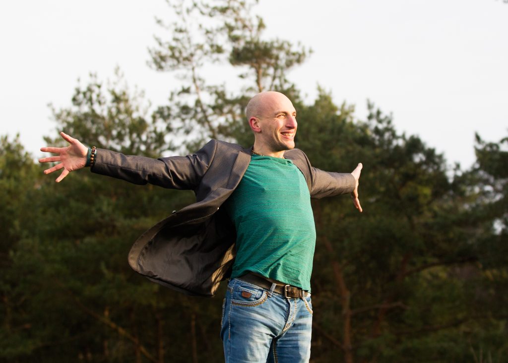 Marco Bot met gespreide armen in de natuur, lachend tussen de dennenbomen