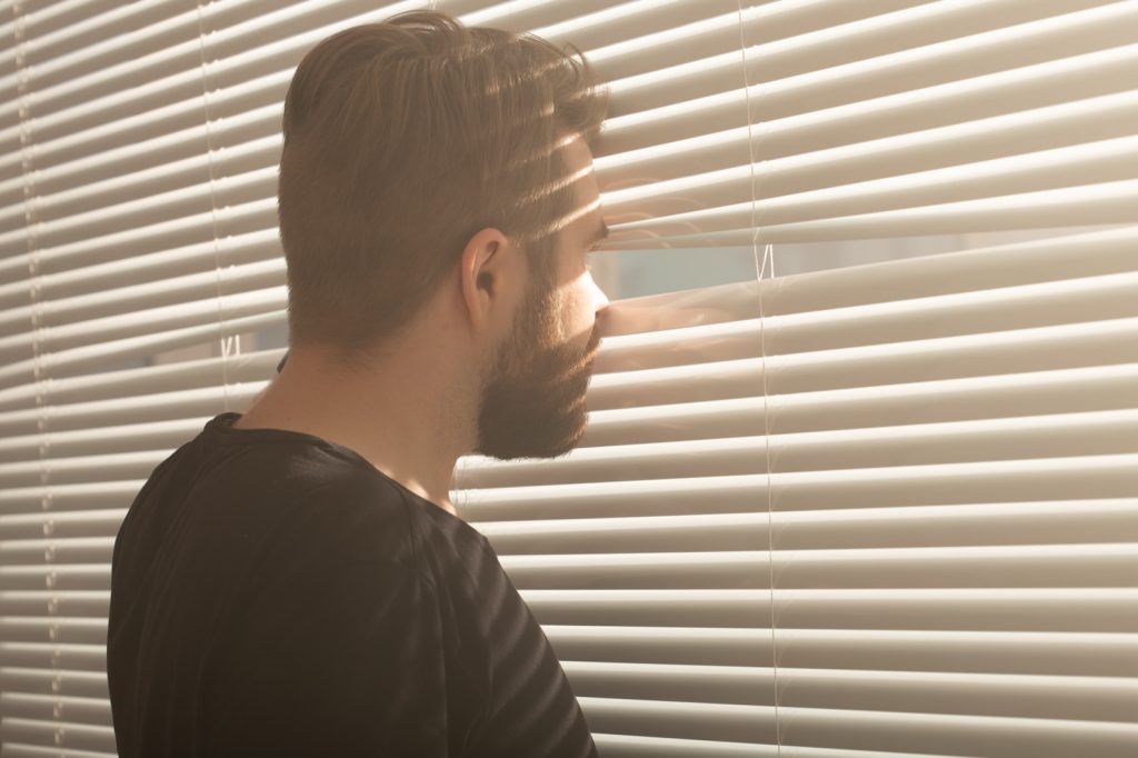 Man met baard kijkt door de lamellen naar buiten, verveeld en afwezig