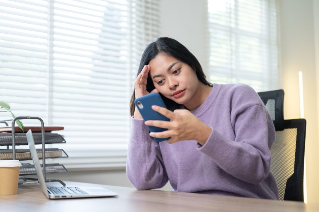 Vrouw aan haar bureau kijkt vermoeid en afwezig naar haar telefoon, met haar hoofd op haar hand